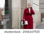 Portrait Stylish Woman in Burgundy Coat in City Street Wearing Fashionable clothing White Turtleneck Sweater, Grey Jeans, Carrying White Bag. Confident and Chic Mature Fashion Model