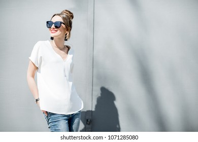 Portrait of a stylish woman dressed in white shirt with sunglasses on the grey wall background outdoors