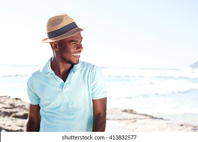 Portrait of stylish young african man with hat looking away and smiling at the beach in summer