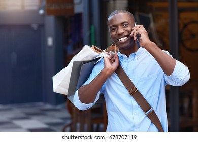 Portrait Of A Stylish Young African Man Smiling And Talking On His Cellphone While Carrying Paperbags While Out Shopping In The City