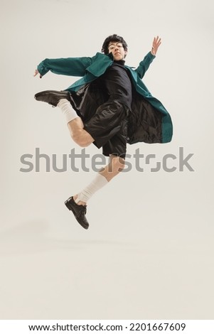 Portrait of stylish young man in black outfit and green coat posing isolated over grey studio background. Looks delightful. Concept of modern fashion, art photography, style, queer, uniqueness, ad