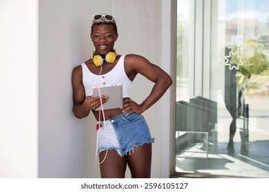 Portrait of stylish young non-binary person using tablet and wearing headphones and sunglasses, looking at camera