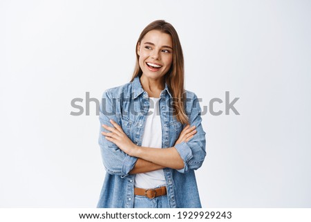 Portrait of stylish young woman with blond hair, cross arms on chest, looking left at empty space for banner and smiling pleased, checking out advertisement, white background