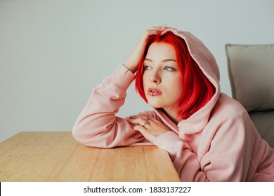 Portrait of a stylish young woman  with bright red hair, lip piercing and hoodie holding head on desk looking  off