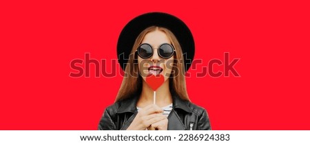 Portrait of stylish young woman with heart shaped lollipop posing wearing black round hat on red background