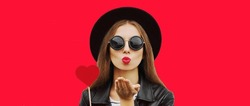 Portrait Of Stylish Young Woman With Red Heart Shaped Lollipop Blowing Her Lips Sending Sweet Air Kiss Wearing A Black Round Hat On Background
