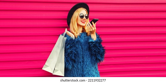 Portrait Of Stylish Young Woman With Shopping Bags And Smartphone Using Voice Command Recorder, Assistant Or Takes Calling In City Wearing Blue Fur Coat, Black Round Hat On Pink Background