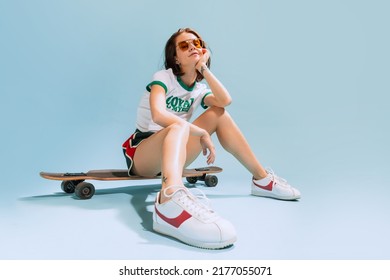 Portrait of stylish young woman sitting on skateboard, posing isolated over blue studio background. Retro fashion outfit. Summertime mood. Concept of beauty, style, emotions, fashion, lifestyle - Powered by Shutterstock