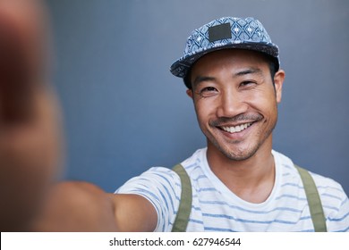 Portrait of a stylishly dressed young Asian man smiling and taking a selfie while standing alone in front of a gray wall outside
