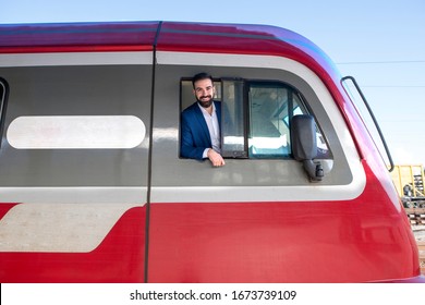 Portrait Of Subway Driver Leaned Out Of His Train Window Cockpit At The Station Before Departure. Railway Transportation And Engine Driver Occupation.