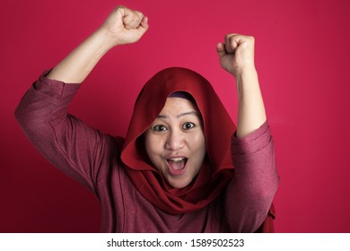 Portrait of success beautiful Asian muslim woman wearing hijab showS winning gesture, excited happy expression celebrating victory, against red background