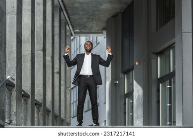 Portrait of successful african american businessman boss, man celebrating victory and success holding hands up dancing office building bells, satisfied employee with achievement results. - Powered by Shutterstock