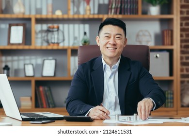 Portrait of a successful Asian banker, businessman working in the office, looking at the camera and smiling, counting metal coins on the table - Shutterstock ID 2124611531
