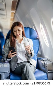 Portrait Of A Successful Asian Business Woman Or Female Entrepreneur In Formal Suit In A Plane Sits In A Business Class Seat And Uses A Smartphone With Drink Coffee During Flight, Relax Concept