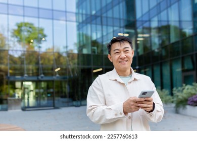 Portrait of successful Asian businessman in casual clothes, man with phone in hands smiling and looking at camera, business owner outside office building, satisfied freelancer startup entrepreneur. - Powered by Shutterstock