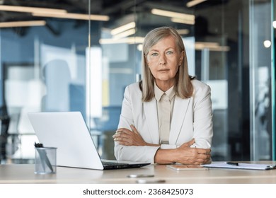 Portrait of a successful beautiful senior business woman sitting in a business suit at a desk, in a company office and looking smug, and serious at the camera. - Powered by Shutterstock