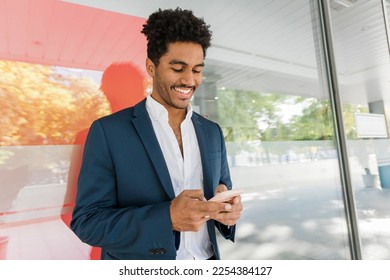 Portrait of successful black businessman in suit leaning on window using Smartphone. Young businessman using mobile phone outside office building