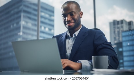 Portrait Of Successful Black Businessman In Tailored Suit Working On Laptop Computer In His Big City Office. Digital Entrepreneur Does Data Analysis, Cloud Computing For E-Commerce Strategy Assessment