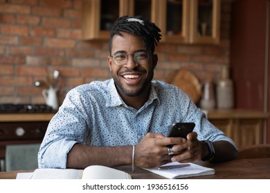 Portrait of successful black male modern day student distracted from working learning look at camera hold smartphone. Happy smiling young afro american man hipster posing at kitchen table do paperwork