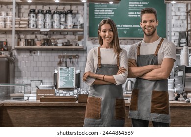 Portrait of successful business coffee shop owners indoors. Photo of young caucasian waiter and waitress standing in uniform with arms crossed and smiling at camera with counter on background - Powered by Shutterstock