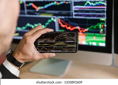 Portrait of successful businessman 30s wearing suit using smartphone while working with digital graphics on computer in office - Shutterstock ID 1333892609