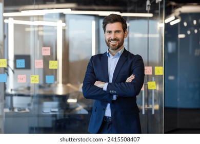 Portrait of successful businessman inside office in business suit, man with crossed arms smiling and looking at camera, experienced financier boss near window, investor.