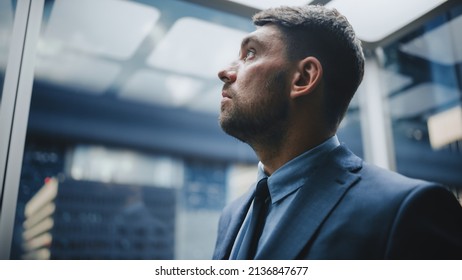 Portrait of Successful Businessman in a Suit Riding Glass Elevator to Office in Modern Business Center. Young Male Looking at Modern Downtown Skyscrapers Out of the Panorama Window in the Lift. - Powered by Shutterstock