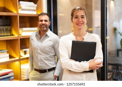 Portrait of successful businesswoman standing with folder in her office, mid adult male colleague standing and smiling behind. Female leader concept