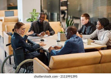 Portrait of successful businesswoman using wheelchair at meeting table and talking to diverse group of colleagues in modern office space