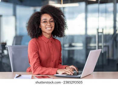 Portrait of successful businesswoman at workplace inside office, woman smiles and looks at camera, uses laptop at work, female programmer in red shirt with curly hair, codes new software. - Powered by Shutterstock