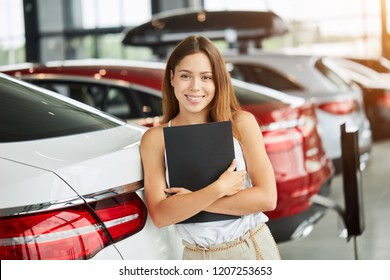 Portrait Of Successful Caucasian Female Sales Representative In Car Dealership. Rental Vehicle Concept. Beautiful Female Seller Or Saleswoman Holding Black Folder Looking At Camera Ready To Help.