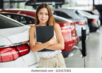 Portrait Of Successful Caucasian Female Sales Representative In Car Dealership. Rental Vehicle Concept. Beautiful Female Seller Or Saleswoman Holding Black Folder Looking At Camera Ready To Help.