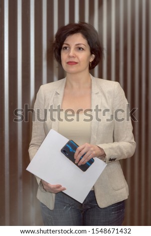 Portrait of successful confidence smiling business woman holding papers with copy space and smart phone. Attractive middle aged woman with beautiful smile in business suit against striped wall.
