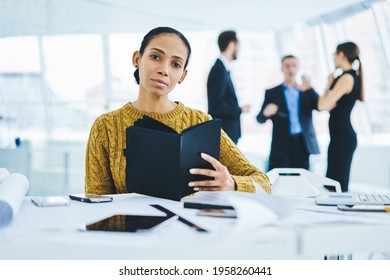 Portrait Of Successful Female Finance Director Looking At Camera While Business Partners On Background Analyzing Strategy,experienced Woman With Black Textbook Planning Trade During Work Day In Office