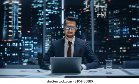 Portrait of a Successful Handsome Businessman Working on Laptop Computer in Big City Office Late in the Evening. Happy Competent Manager in Stylish Dark Blue Suit Smiles and Poses for Camera.