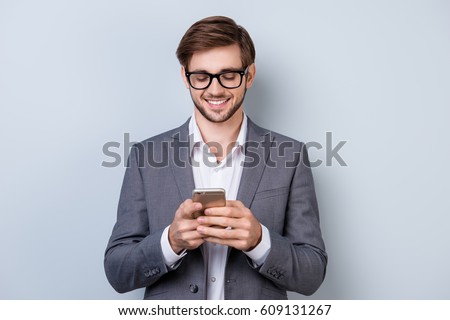 Portrait of successful handsome young man in formal wear with beaming smile, bristle and glasses holding smartphone and read sms while standing on gray background.