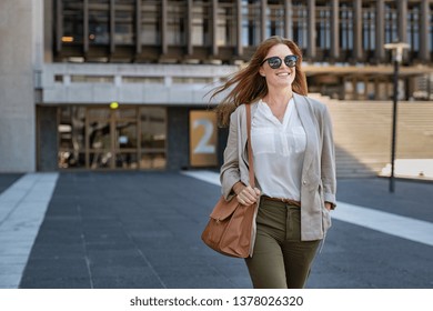 Portrait of successful happy woman on her way to work on street. Confident business woman wearing blazer carrying side bag walking with a smile. Smiling woman wearing sunglasses and walking on street.
