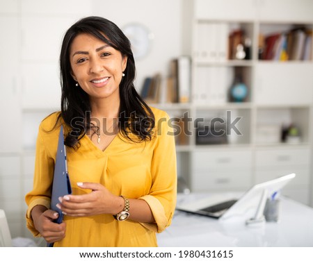 Portrait of successful Hispanic business woman with folder of documents in her hands, standing in modern office