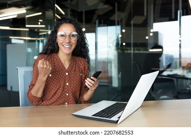 Portrait Of Successful Latin American Business Woman, Worker In Glasses And Curly Hair Looking At Camera And Smiling Happy Celebrating Triumph And Winning Success Sitting At Table Holding Phone