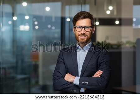 Portrait of successful mature boss, senior businessman in glasses and business suit looking at camera and smiling, man with crossed arms working inside modern office building