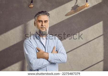 Portrait of a successful mature businessman. Bearded grey haired senior business man in a blue shirt standing with his arms folded against a grey office wall background