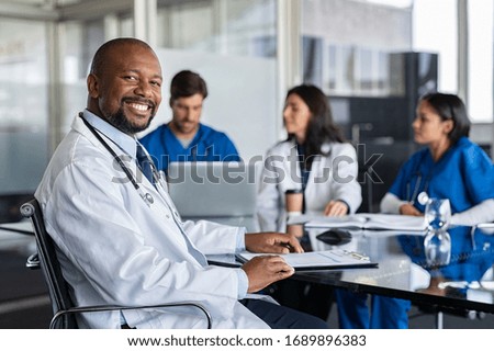 Portrait of successful mature doctor looking at camera with working clinicians behind. Smiling african clinic worker in labcoat and stethoscope sitting in conference room with copy space in hospital.