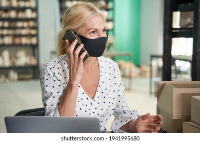 Portrait of successful mature woman, small business owner wearing protective mask looking away while making a call, sitting at the desk in her craft pottery shop - Powered by Shutterstock