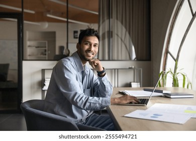 Portrait of successful millennial Indian male entrepreneur posing for camera seated in cozy workspace with wireless computer. Productivity, competence, workflow using modern technology and internet - Powered by Shutterstock