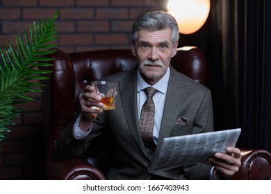 Portrait Of Successful Millionaire And Mature Handsome Old Man Sitting In Leather Chair Of His Office With Whiskey And Newspaper In His Hands.