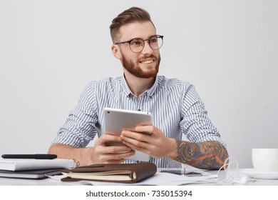 Portrait Of Successful Office Worker In Round Spectacles, Has Tattooed Arms, Holds Modern Tablet, Makes Annual Report, Going To Have Minute Rest And Surf Social Networks. Technolgy, Business Concept