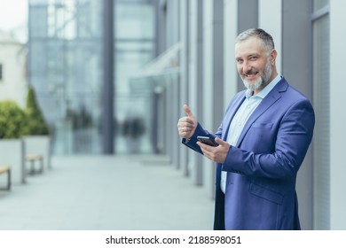 Portrait Of Successful Senior Gray Haired Banker, Arab Man Looking At Camera Smiling And Holding Finger Up, Businessman Outside Office Using Smartphone