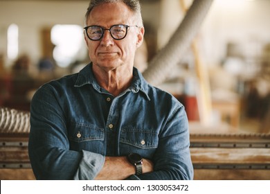 Portrait of successful senior man in eyeglasses standing in his carpentry workshop. Proud carpentry workshop owner standing with his arms crossed. - Powered by Shutterstock