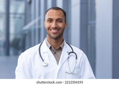 Portrait Of Successful And Smiling African American Doctor, Man In Medical Coat With Stethoscope Looking At Camera And Smiling, Outside Modern Hospital