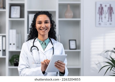 Portrait of successful smiling hispanic female doctor inside medical office, woman with tablet computer close up smiling and looking at camera, working in medical gown while standing. - Powered by Shutterstock